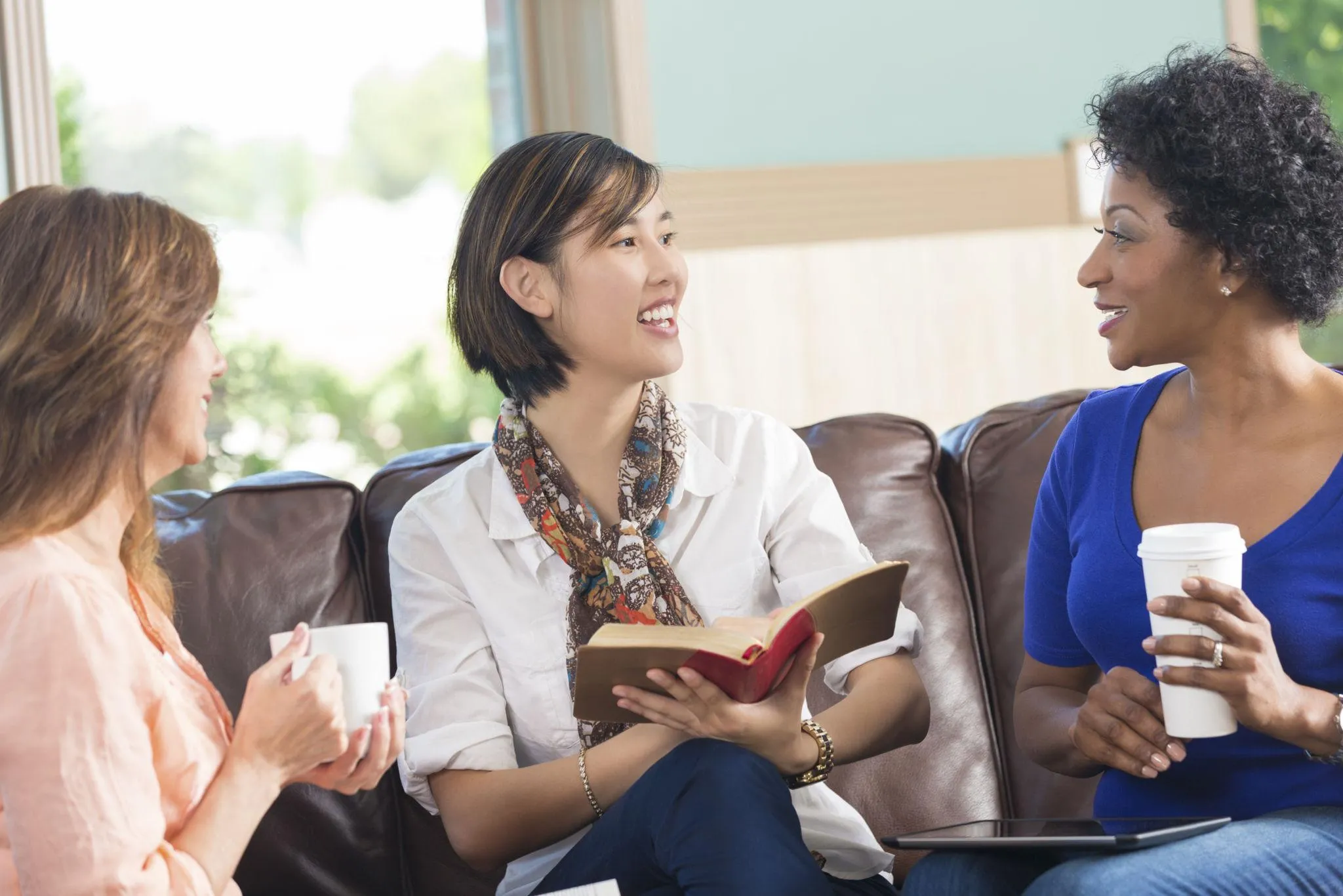 Three women sitting on a leather sofa, holding coffee mugs, laughing and talking during a Bible study session Three women sitting on a leather sofa, holding coffee mugs, laughing and talking during a Bible study session